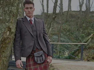 Kilt Outfits to buy. Medium Close view of a man wearing traditional highlandwear including a jacket, tie that matches his kilt tartan, waistcoat, and leather sporran. He is standing, leaning on a tree, facing camera.