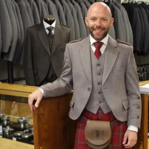 Bold, smiling man in full kilt outfit standing in front of a wooden counter of a kilt hire shop, with an assortment of jackets for hire hanging behind him. Also offering made to measure kilts.